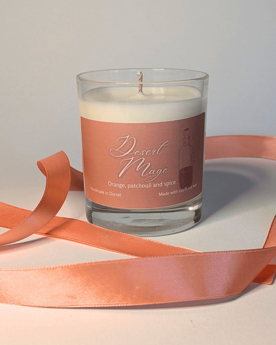a soy candle with a pale orange label, reading 'Desert Mage'. the table in front of the candle is decorated with a ribbon in the same orange shade.