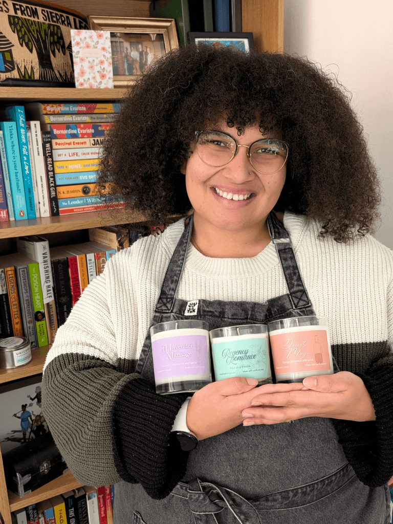 a young black woman holds three candles in her hands. she smiles at the camera, standing in front of a bookshelf filled with books.